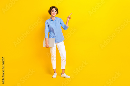 Full size photo of cheerful woman bob hairdo dressed blue blouse indicating empty space hold laptop isolated on yellow color background