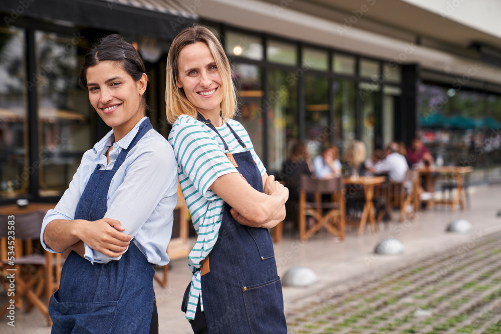 Photo of two happy female restaurant owners standing back to back in front of their restaurant