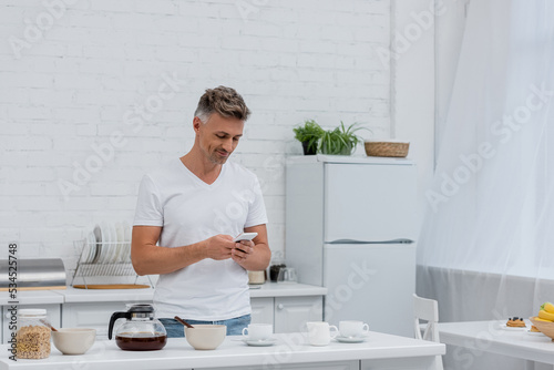 Smiling man using smartphone near breakfast and coffee pot in kitchen.