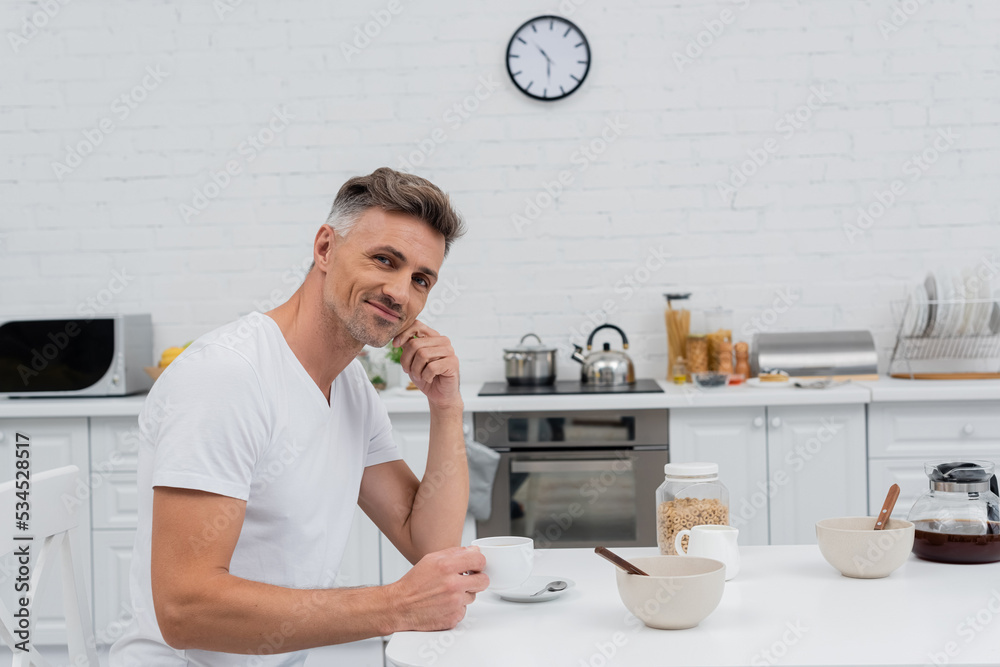 Smiling man holding cup of coffee and looking at camera near cereal rings in kitchen. Smiling man holding cup of coffee and looking at camera near cereal rings in kitchen.