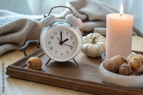 White vintage alarm clock, candle, autumn decoration and sweets on a nightstand with a natural woolen blanket, fall back concept after daylight saving time