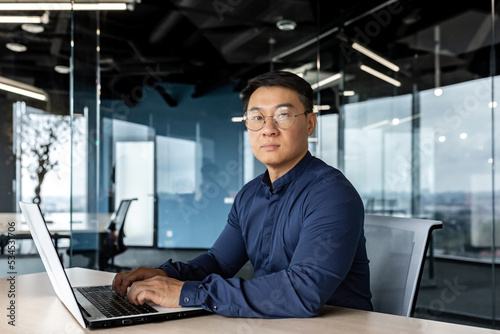 Serious and focused businessman looking at camera, asian man working inside modern building, man wearing glasses and casual shirt using laptop. © Liubomir