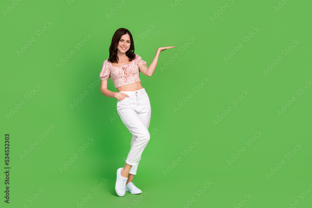 Full size photo of incredible girl dressed blouse white pants hand in pocket demonstrating product isolated on green color background Full size photo of incredible girl dressed blouse white pants hand in pocket demonstrating product isolated on green color background