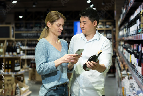 Dissatisfied international couple, Asian man and woman buying wine. They are standing in the alcohol department in the supermarket, unable to choose a wine, holding a bottle and a phone, arguing.