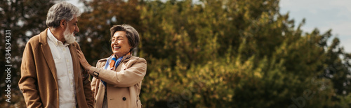 joyful senior woman hugging bearded husband in coat and standing in park, banner. © LIGHTFIELD STUDIOS