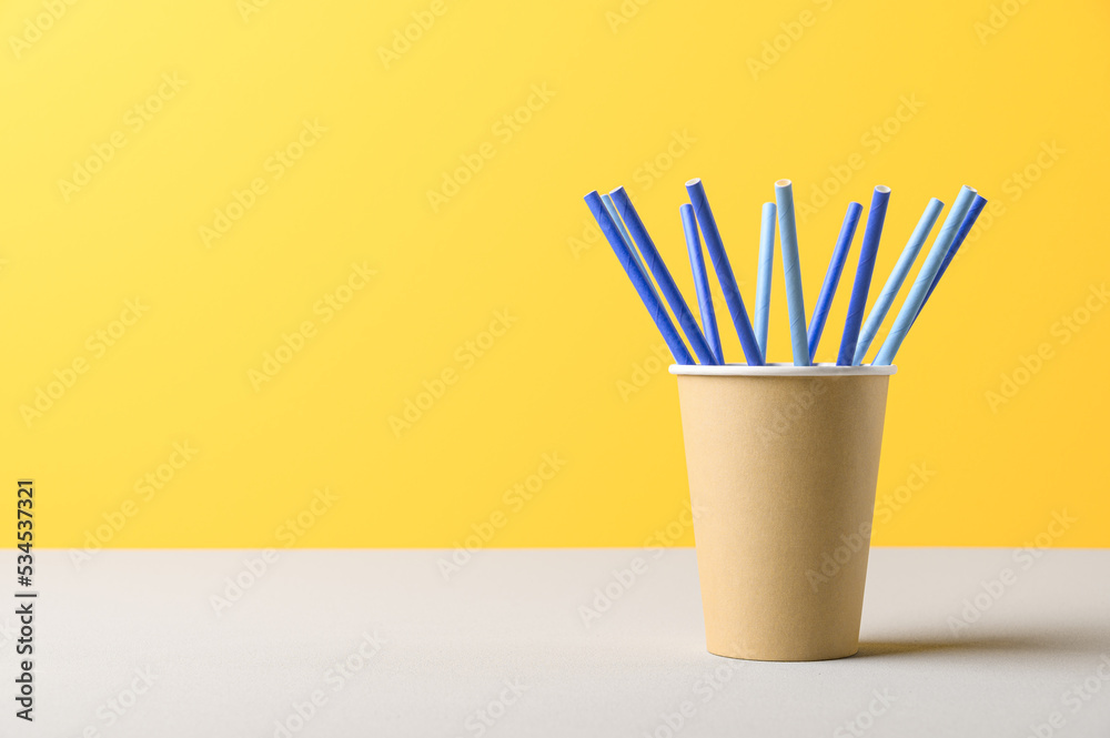 Pile of blue straws in craft paper cup for birthday party. Bright yellow wall. Pop Art style. Recycling eco concept. Space for text Pile of blue straws in craft paper cup for birthday party. Bright yellow wall. Pop Art style. Recycling eco concept. Space for text