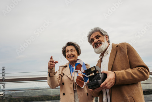 bearded and senior man holding vintage camera near wife smiling while pointing with finger on bridge.