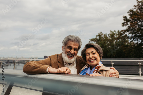 bearded senior man in coat hugging cheerful wife and standing near bridge guard rail.