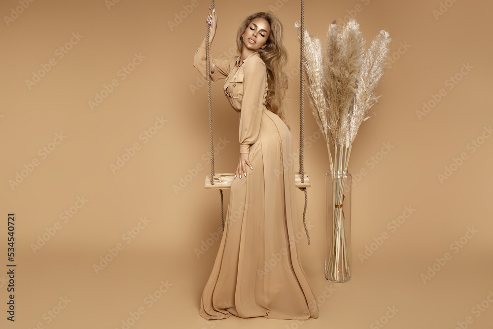 Autumn fashion. Elegant woman in beige long dress is swinging on wooden swing on beige background in studio.