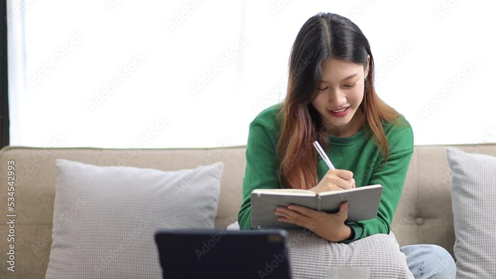 Charming Asian woman in the living room at home reading and writing in a diary book. Charming Asian woman in the living room at home reading and writing in a diary book.
