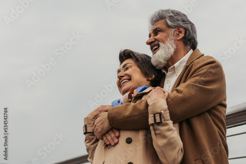 low angle view of happy senior man hugging positive wife in beige coat and smiling outside.
