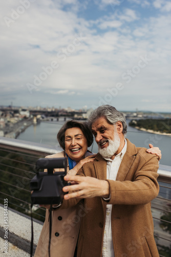 happy senior man taking selfie with cheerful wife on vintage camera with blurred city on background.