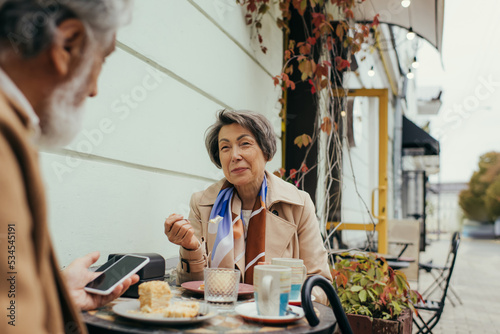 happy senior woman eating cake and looking at husband with smartphone on terrace of cafe.