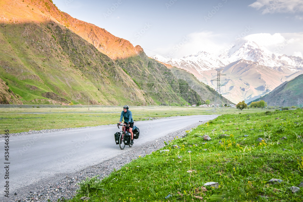 Caucasian male cyclist bicycle touring in caucasus KAzbegi mountains and Kazbek peak in the background. Travel around caucasus. Cycle around the world Caucasian male cyclist bicycle touring in caucasus KAzbegi mountains and Kazbek peak in the background. Travel around caucasus. Cycle around the world