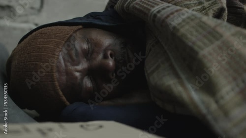 Close up shot of African American homeless man in knitted hat sleeping in rags on ground