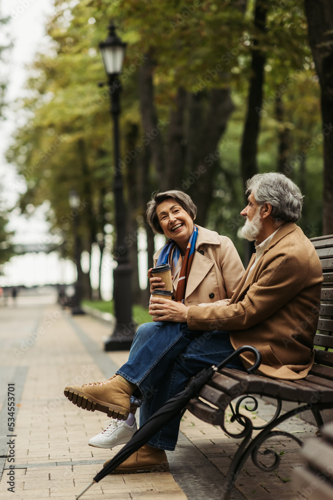 full length of happy senior couple in coats holding paper cups while sitting on bench in park.