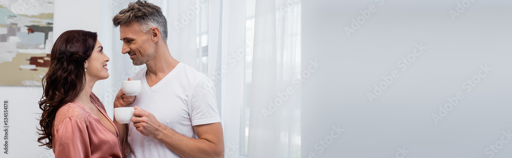 Side view of smiling brunette woman in silk robe looking at husband with coffee cup at home, banner. Side view of smiling brunette woman in silk robe looking at husband with coffee cup at home, banner.