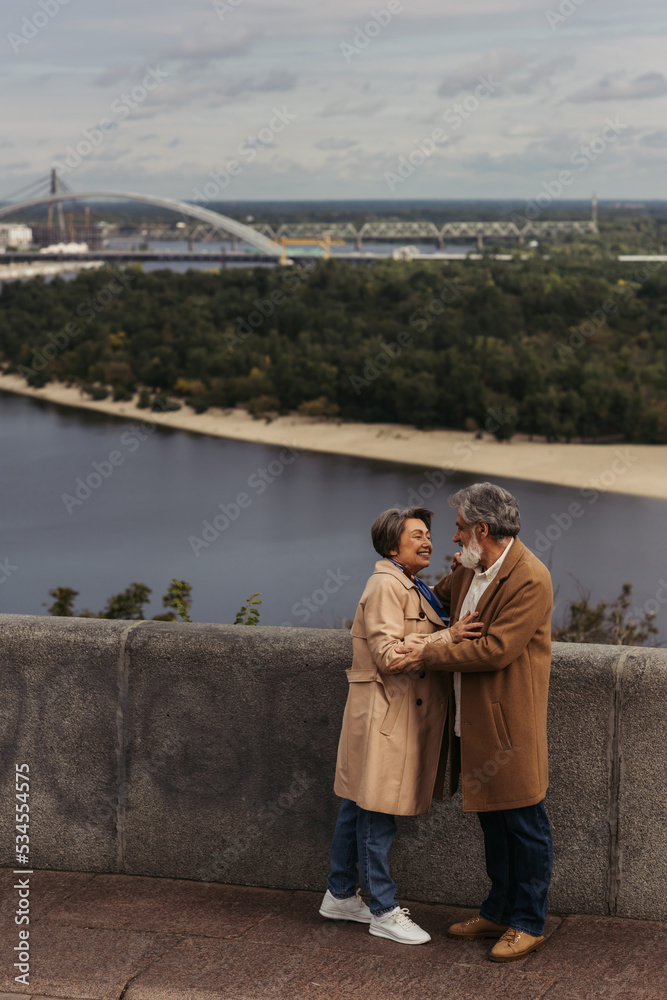 full length of happy senior couple in beige autumnal coats hugging on bridge near river.