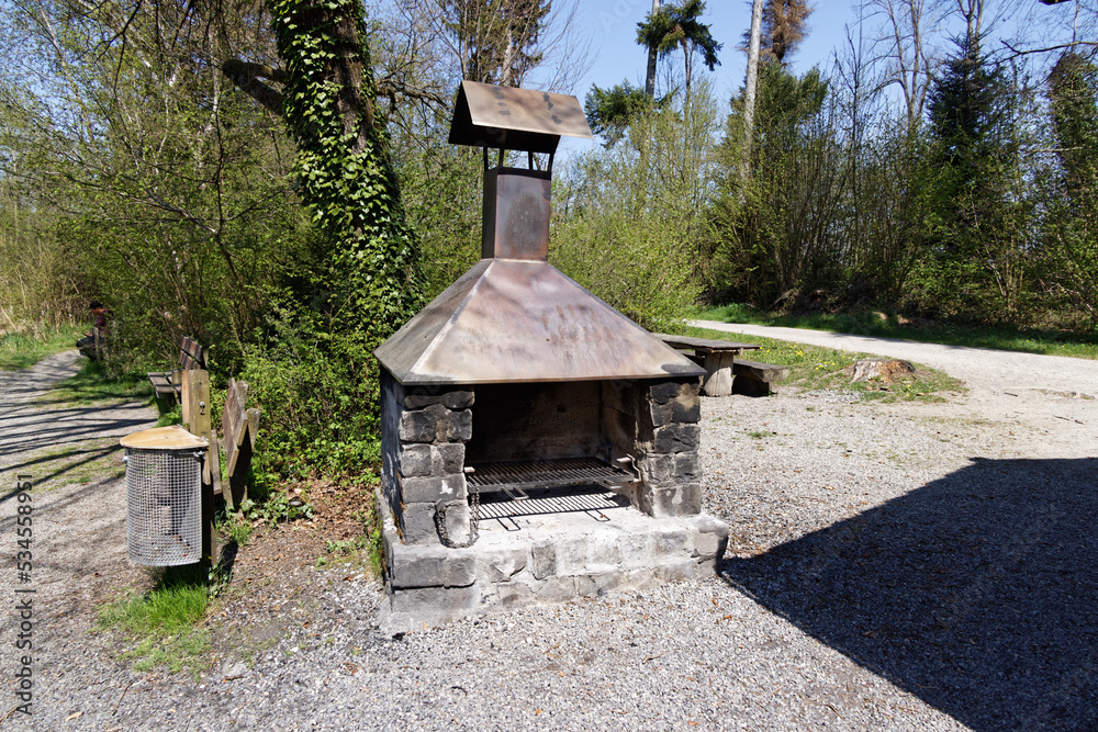 brick chemine oven with copper roof, metal grill without meat, the grill is not in use, day,sunshine, tree with ivy, gravel parking and lattice wastebasket