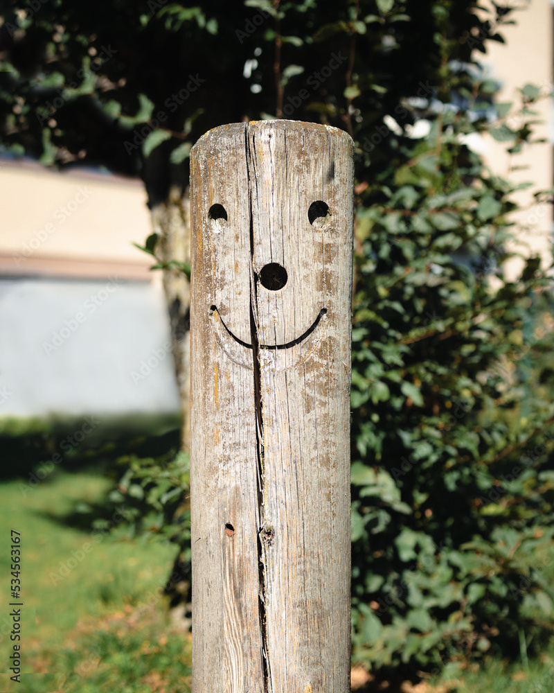 Cute smiley face carved in wood pole outside in a playground. Happy and funny face carving as decoration on fence post. Lucerne Switzerland. Selective focus with defocused plants and building. Cute smiley face carved in wood pole outside in a playground. Happy and funny face carving as decoration on fence post. Lucerne Switzerland. Selective focus with defocused plants and building.