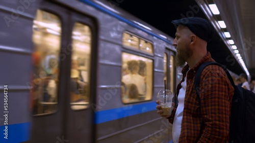 A bearded man in a cap stands on a subway platform and drinks iced coffee while waiting for a subway train. A typical day on the subway, a man goes to work every day early in the morning.