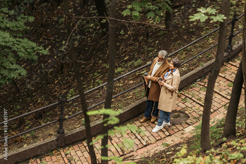 high angle view of cheerful and retired couple in beige coats holding paper cups while walking in park.