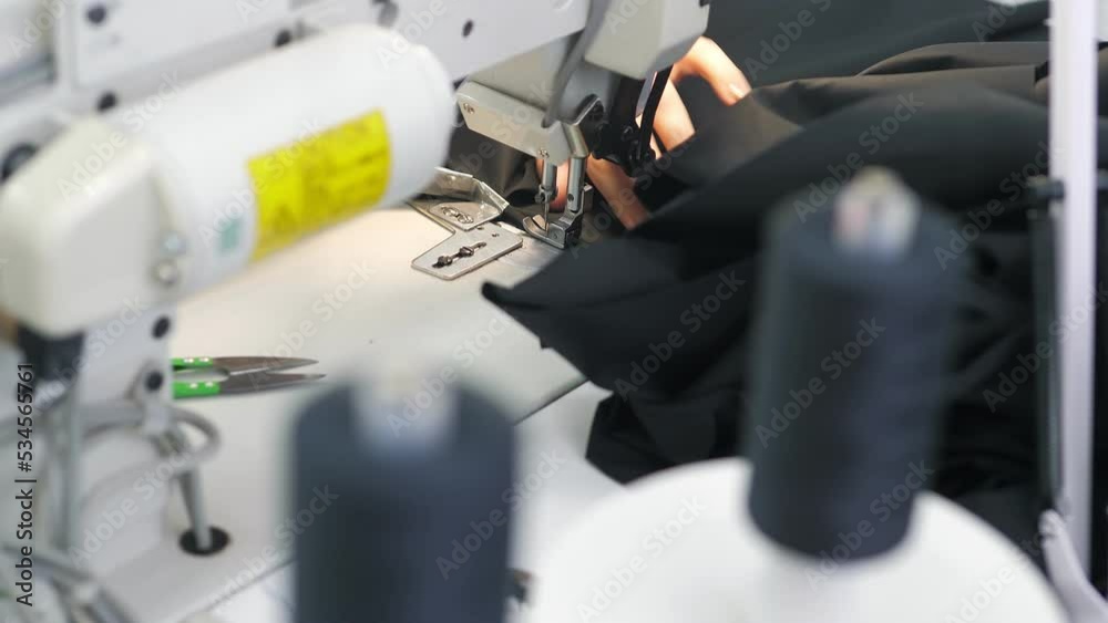 Woman sews black fabric on professional sewing machine while sitting at her working place in fashion workshop.The seamstress's hands hold textiles for the production of clothes.Designer sew the dress Woman sews black fabric on professional sewing machine while sitting at her working place in fashion workshop.The seamstress's hands hold textiles for the production of clothes.Designer sew the dress