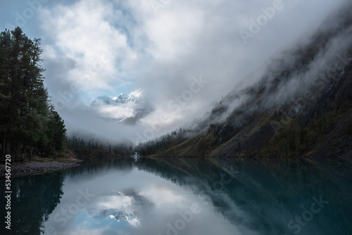 Tranquil scenery with snow castle in clouds. Mountain creek flows from forest hills into glacial lake. Snowy mountains in fog clearance  small river and coniferous trees reflected in calm alpine lake.