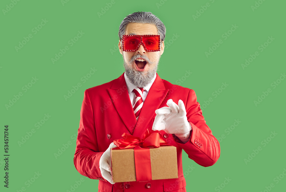 Wondered cheerful emotional old man with opened mouth looking at camera, showman wearing in Santa style holding Christmas gift in hands on green background. Celebrating Christmas, New Year concept. Wondered cheerful emotional old man with opened mouth looking at camera, showman wearing in Santa style holding Christmas gift in hands on green background. Celebrating Christmas, New Year concept.
