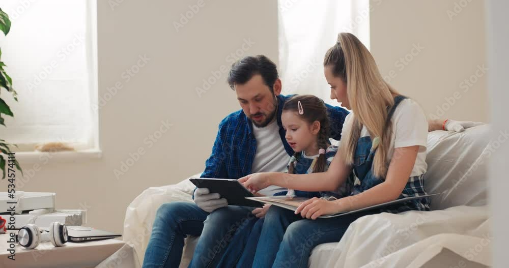 Blond mother father and 8-year-old cute daughter are very happy that their house has been renovated. A woman holds a folder with a color palette on lap, and a man holds a tablet in right hand.