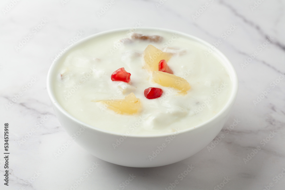 Close-up of Fruits raita or Yogurt & Pineapple or Ananas Raita garnished with pineapple piece and pomegranate, served in a white ceramic bowl over whiet granite background Close-up of Fruits raita or Yogurt & Pineapple or Ananas Raita garnished with pineapple piece and pomegranate, served in a white ceramic bowl over whiet granite background