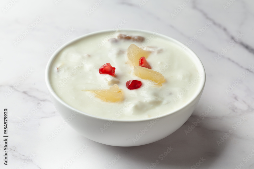 Close-up of Fruits raita or Yogurt & Pineapple or Ananas Raita garnished with pineapple piece and pomegranate, served in a white ceramic bowl over whiet granite background Close-up of Fruits raita or Yogurt & Pineapple or Ananas Raita garnished with pineapple piece and pomegranate, served in a white ceramic bowl over whiet granite background