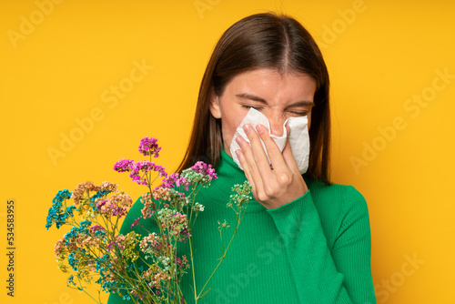 Woman allergic to wild flowers blowing nose in handkerchief with closed eyes on yellow background