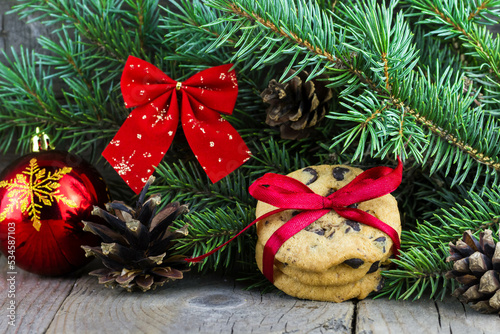 Christmas (New Year) decoration composition. Chocolate chip cookies decorated red bow, fur-tree branches, glass ball and cones on wooden background