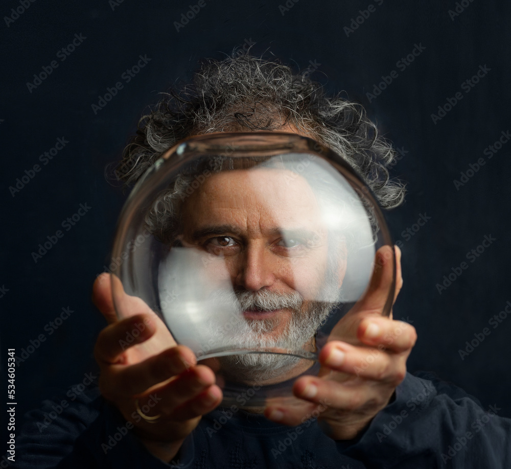 a colorful man with curls, a beard and a silver-colored mustache holds a transparent glass ball in his hands and looks through this ball - obrazy, fototapety, plakaty 
