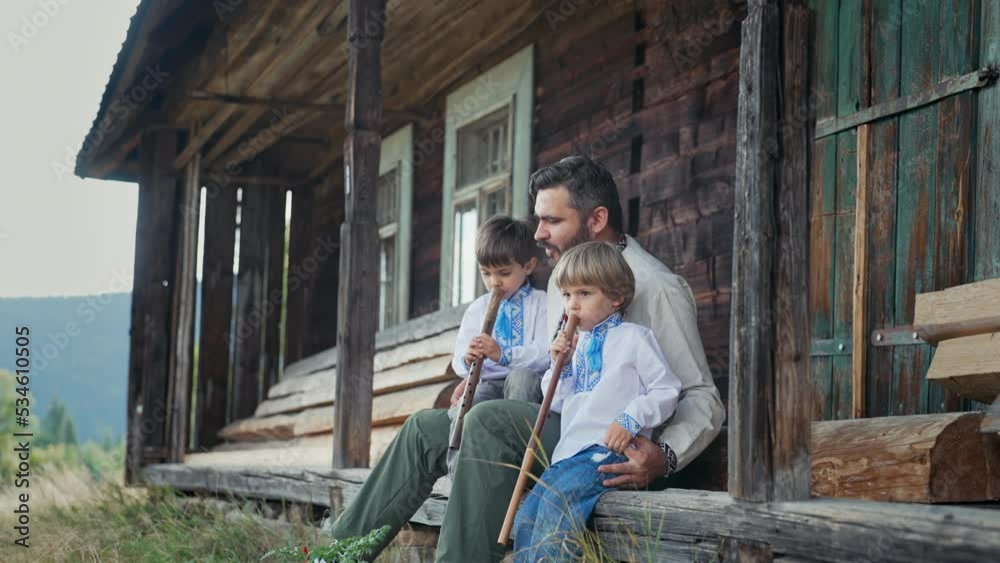 Little boys with dad playing on woodwind wooden flutes - ukrainian sopilka. Family, folk music concept. Father and sons in traditional embroidered shirts. They sitting on porch of old wooden house Little boys with dad playing on woodwind wooden flutes - ukrainian sopilka. Family, folk music concept. Father and sons in traditional embroidered shirts. They sitting on porch of old wooden house