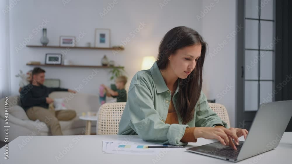 Busy mother working with laptop typing while her small son and girl is playing at home. Business people, family and house concept. Busy mother working with laptop typing while her small son and girl is playing at home. Business people, family and house concept.