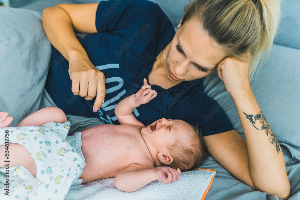 Caucasian young mom laying in bed with her couple months old baby boy. Her son is wearing a diaper, screaming and crying and she is trying to calm him down. High quality photo Caucasian young mom laying in bed with her couple months old baby boy. Her son is wearing a diaper, screaming and crying and she is trying to calm him down. High quality photo