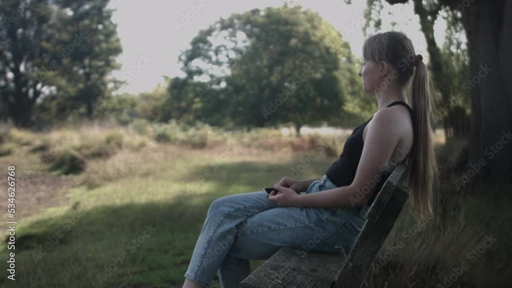 A woman sitting in the bench under the shade of tree and looking out at the distance.