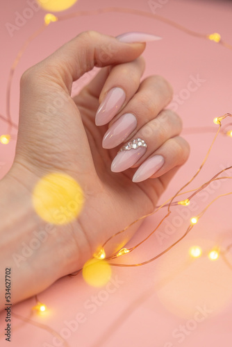 Gorgeous sparkly nude pink beige gel nails with rhinestones on a light pink background with amber LED fairy light string - POV