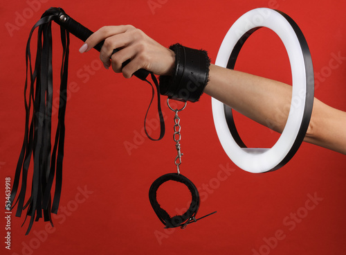 Woman's hand in sex shop leather handcuffs holding whip through led ring lamp on red background. Sex games. Creative idea.