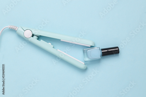 Mini hair straightener with nail polish bottle on a blue background. Pastel color. Top view