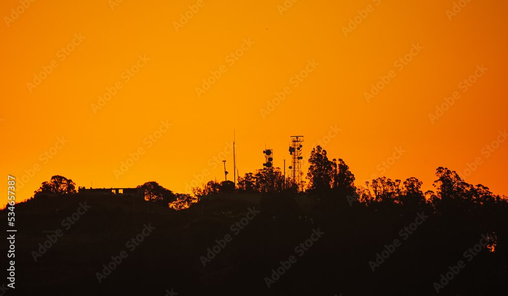 Telecommunication scafolding tower with internet data voice and GSM antennas photographed on top of a hill against amazing orange sunrise sky. Communication technology industry mixed with nature.