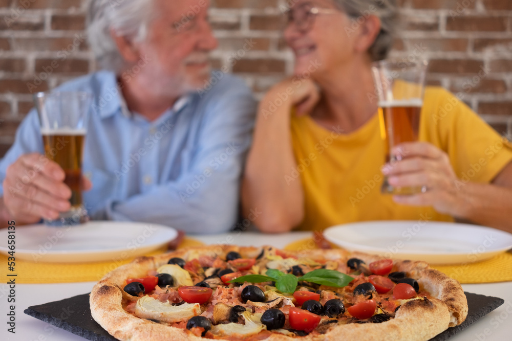 Defocused smiling elderly couple sitting at home table sharing a large pizza, holding two glasses of beer