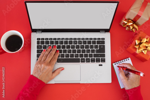 Woman's hands typing on laptop keyboard  at Christmas time, blank space on display for text - red background with Christmas presents