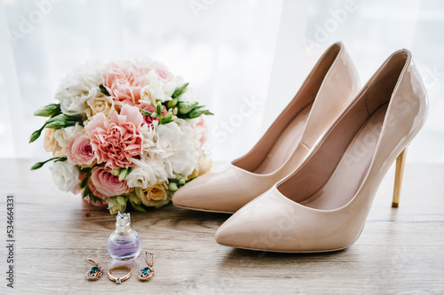 Wedding bouquet of the bride of pink flowers roses and greenery, stylish classic lacquered beige shoes, perfume, earring, and ring lying on wooden background. Bride accessories. Close up. Side view.