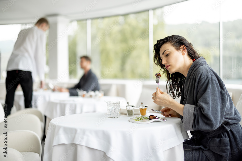Beautiful woman in bathrobe enjoys delicious meal, sitting in white restaurant of haught cuisine during vacations at luxury spa resort Beautiful woman in bathrobe enjoys delicious meal, sitting in white restaurant of haught cuisine during vacations at luxury spa resort