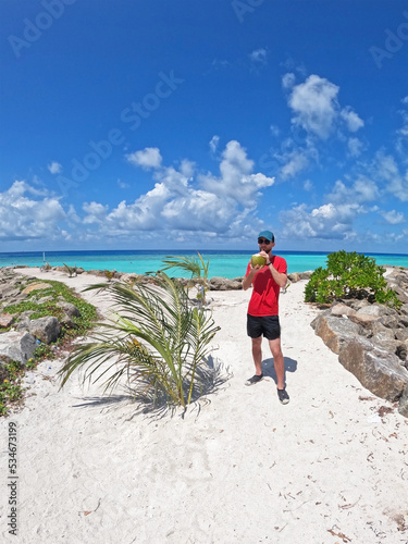 A man drinking fresh coconut juice on a tropical beach against a backdrop of the bright turquoise ocean in the Maldives. Wide angle lens photo