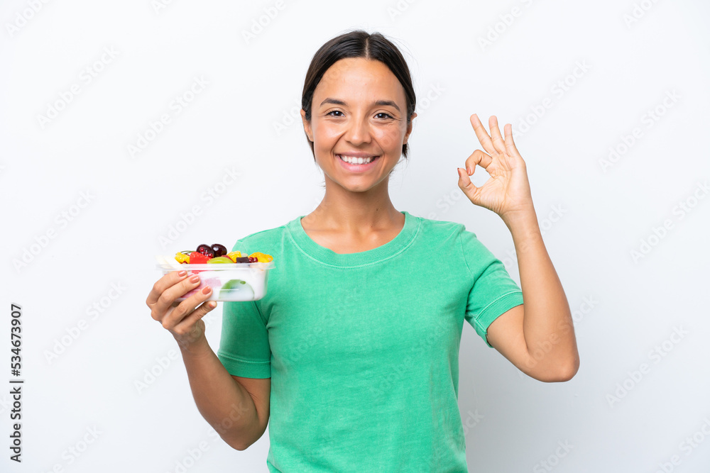 Young hispanic woman holding a bowl of fruit isolated on white background showing ok sign with fingers Young hispanic woman holding a bowl of fruit isolated on white background showing ok sign with fingers