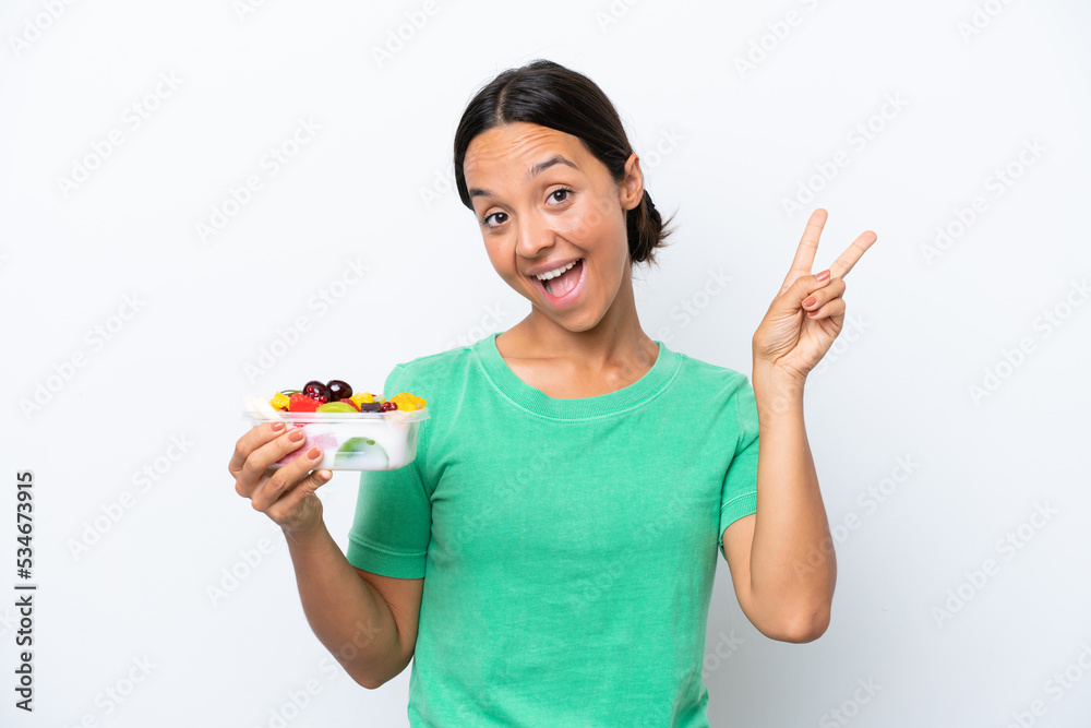 Young hispanic woman holding a bowl of fruit isolated on white background smiling and showing victory sign Young hispanic woman holding a bowl of fruit isolated on white background smiling and showing victory sign